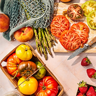 Fresh produce including heirloom tomatoes, peaches, asparagus, and strawberries arranged with a basket and mesh bag, plus sliced tomatoes on a cutting board with a knife.